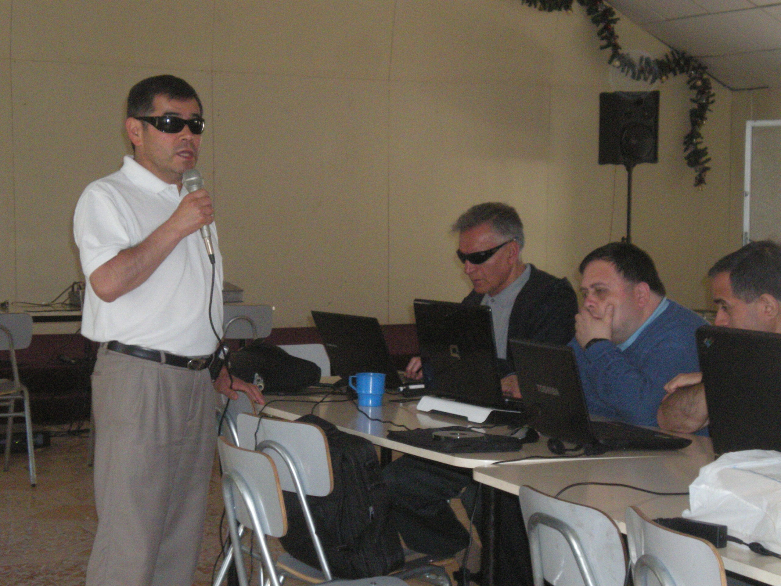 Un hombre habla por un micrófono. Frente a él varias personas sentadas detrás de una mesa usan laptops.
